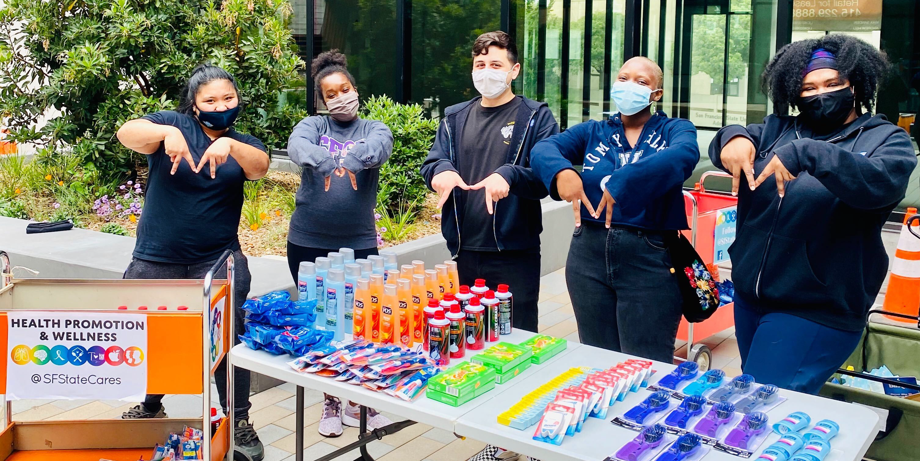 5 individuals wearing dark clothing, demonstrating a hand sign to proudly represent their organization. These individuals are standing behind a table of care and hygiene items to provide to students