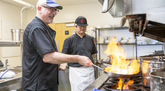 Two male presenting individual cooking with a frypan on fire in an industrial kitchen